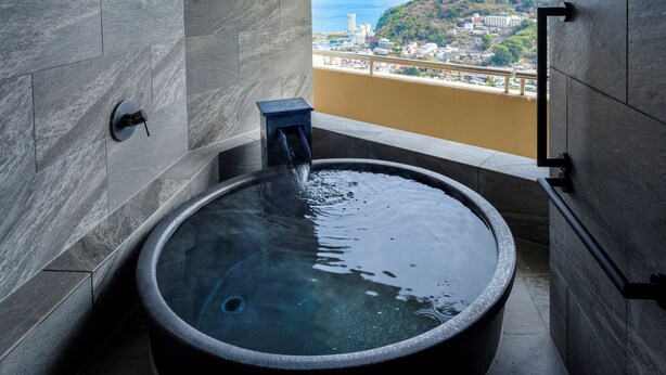 A modern bathroom with a large bathtub, a small stool, and a view of a cityscape and sea. - Kamenoi Hotel Atami (Atami)
