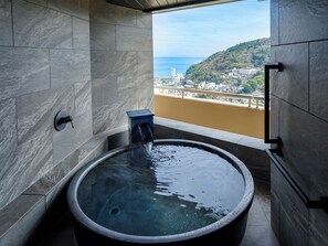 A modern bathroom with a large bathtub, a small stool, and a view of a cityscape and sea. - Kamenoi Hotel Atami (Atami)