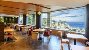 A modern restaurant with high tables and chairs, large windows offering a city view, and a green wall with plants. - Kamenoi Hotel Atami (Atami)
