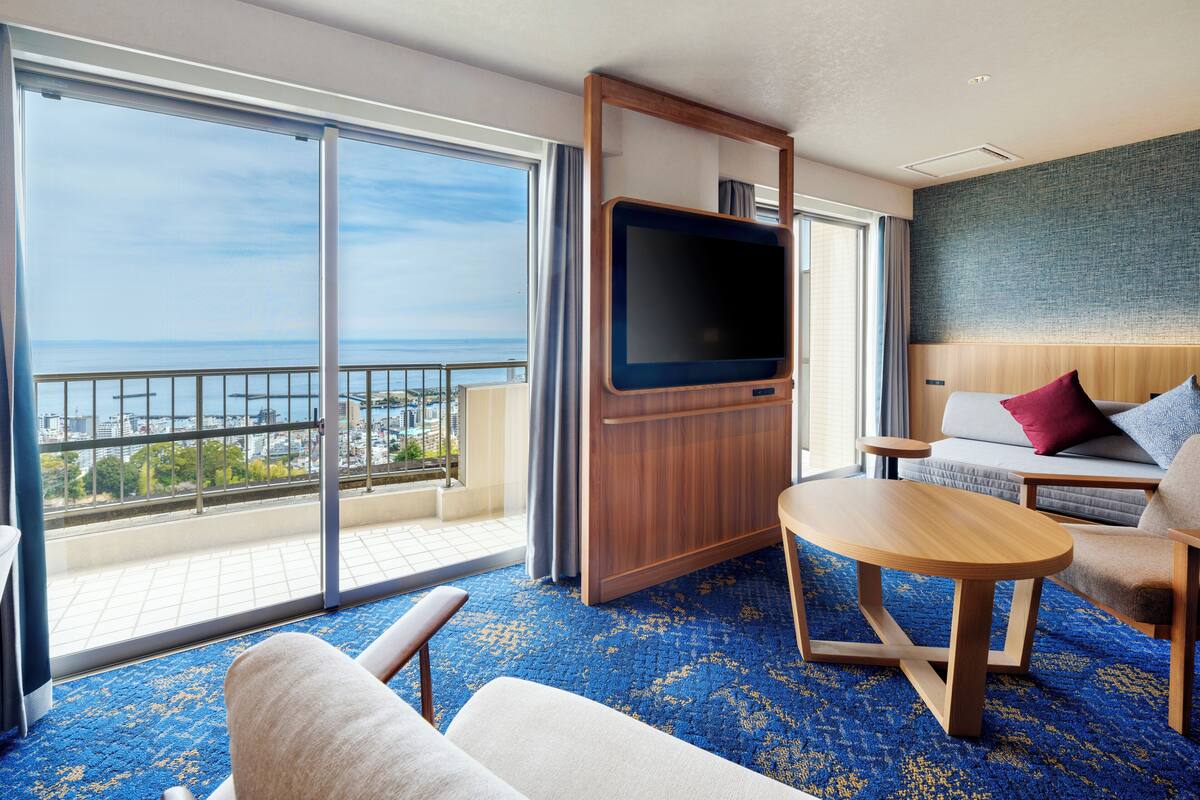 Een moderne hotelkamer met een groot balkon, een flatscreen-tv, een bank, een bijzettafeltje en een kamerbreed tapijt met een patroon.