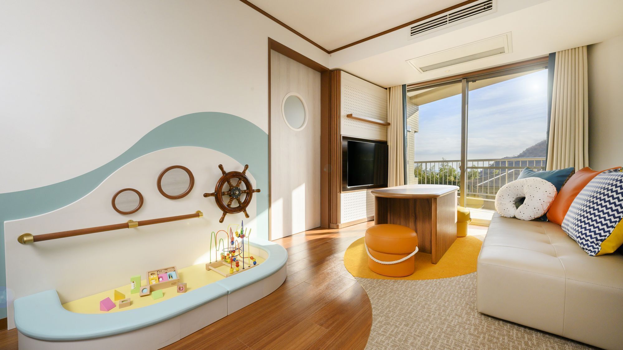 A modern living room with a nautical theme, including a ship's wheel, a small play area, a sofa with cushions, and a large window offering a view.