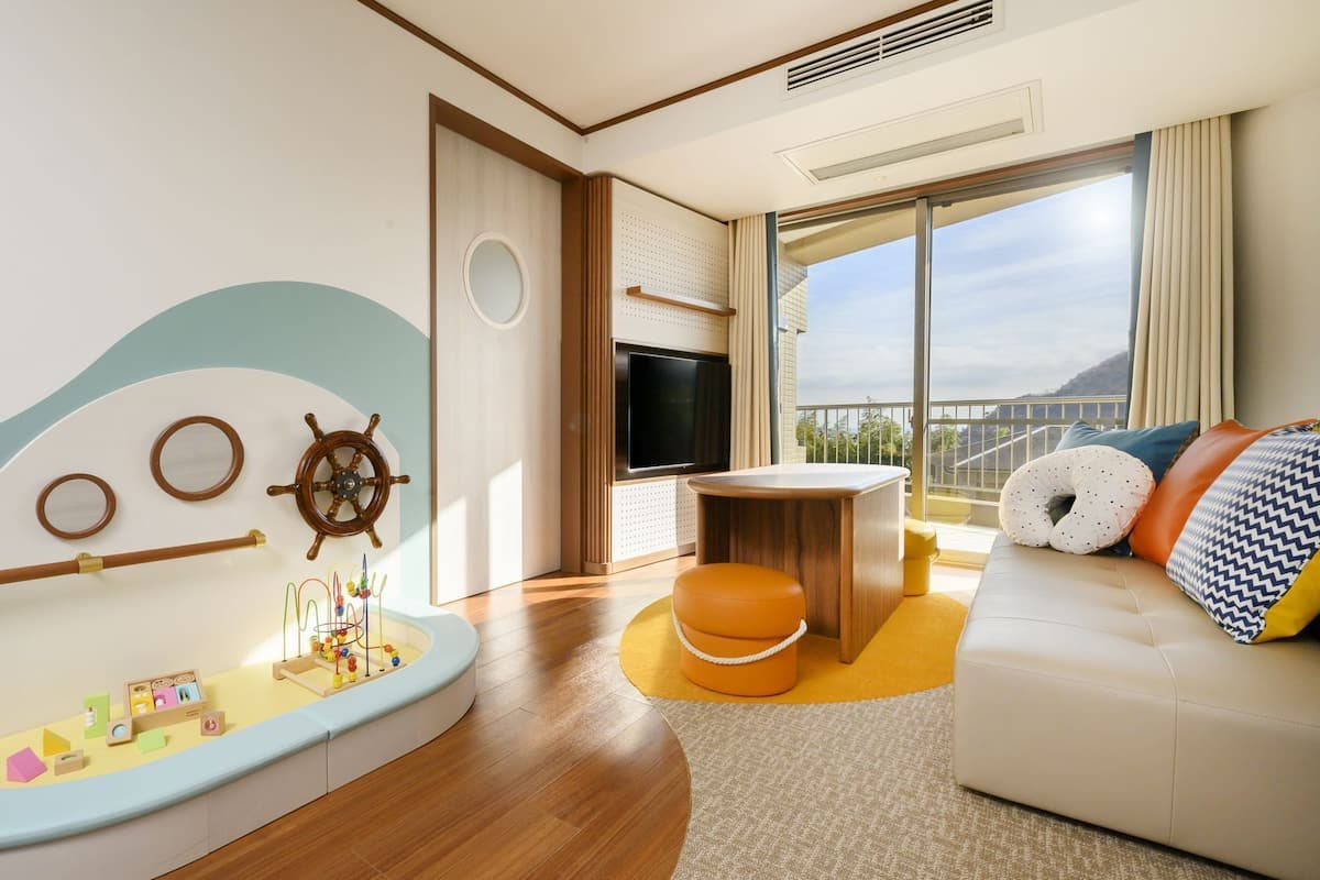 A modern living room with a nautical theme, including a ship's wheel, a small play area, a sofa with cushions, and a large window offering a view.