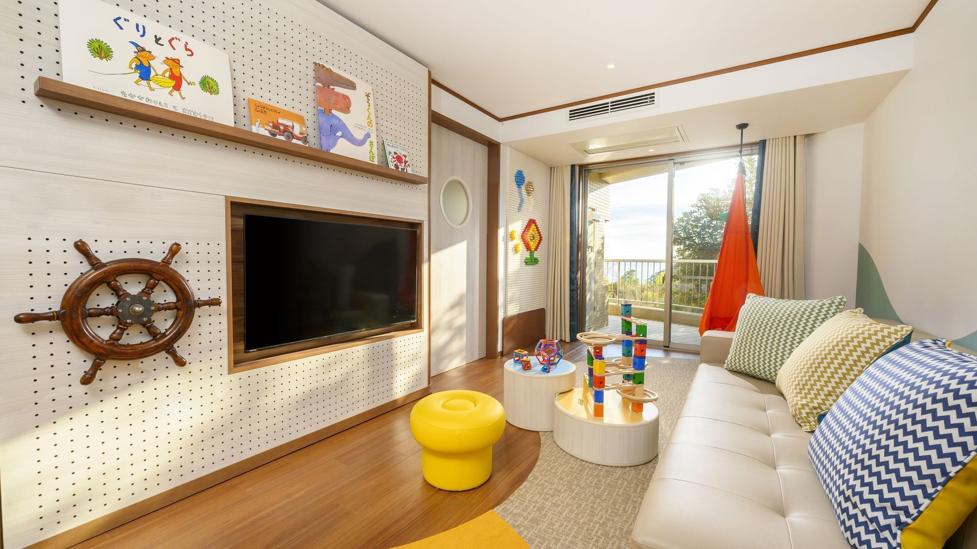 A modern living room with a mounted TV, wooden floor, a yellow ottoman, a white sofa with patterned cushions, and children's toys on a side table.