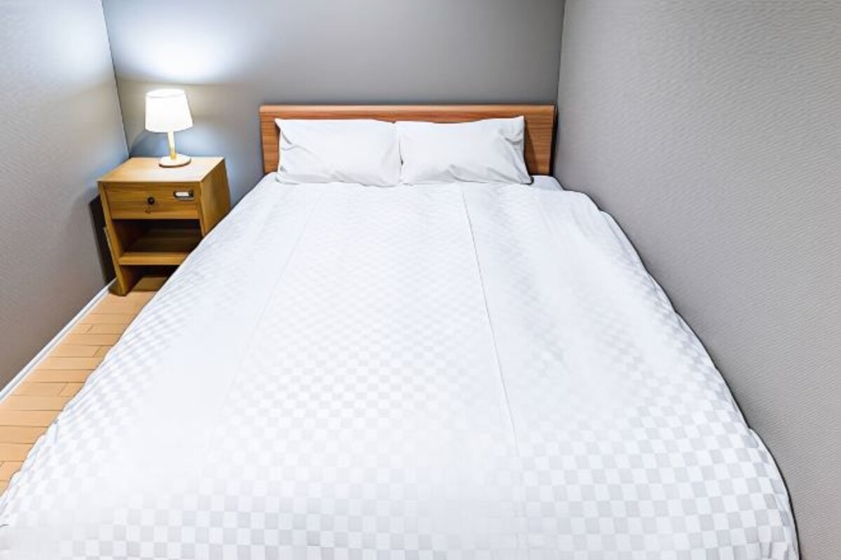 A neatly made bed with white bedding and a wooden headboard, a bedside table with a lamp, and a wooden nightstand.