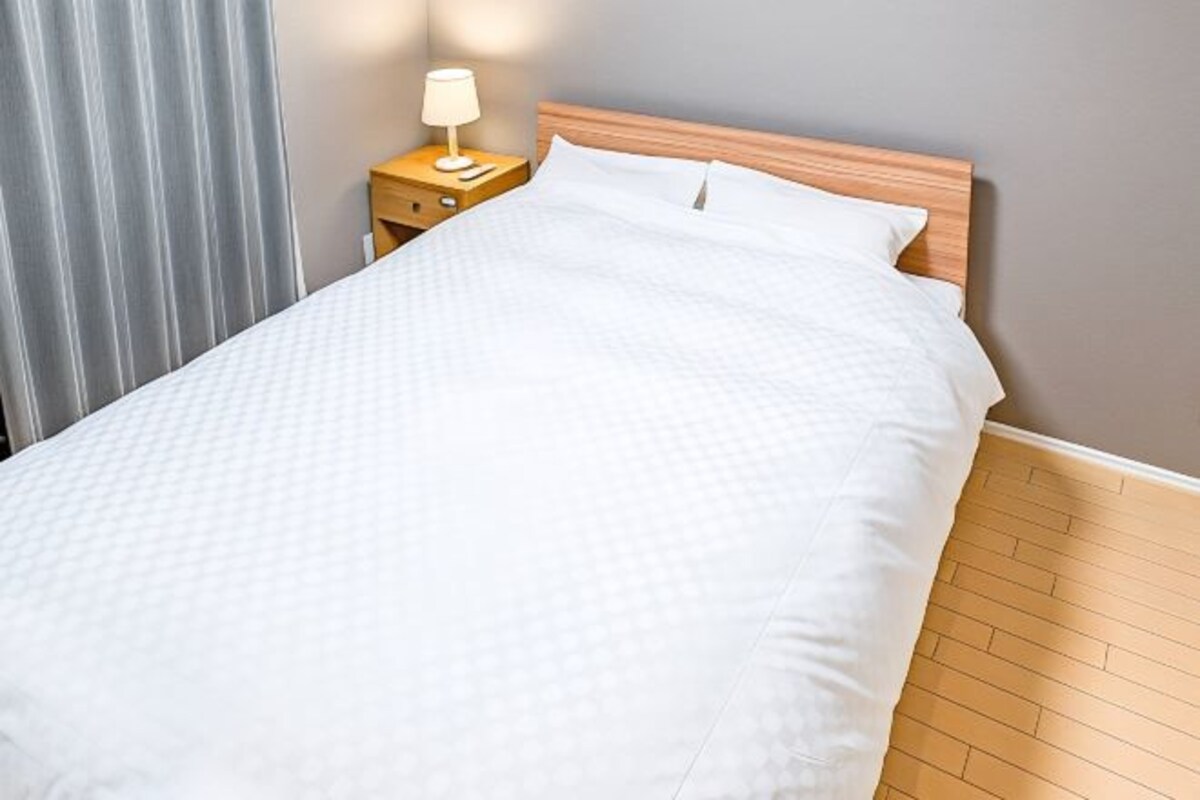 A neatly made bed with a wooden headboard, a bedside table with a lamp, and a grey curtain.