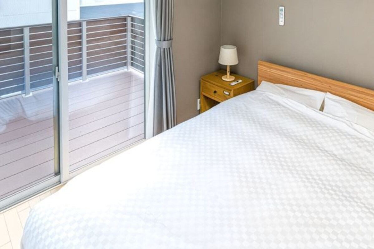 A hotel room with a bed, a nightstand with a lamp, a view of a balcony, and a wall switch.