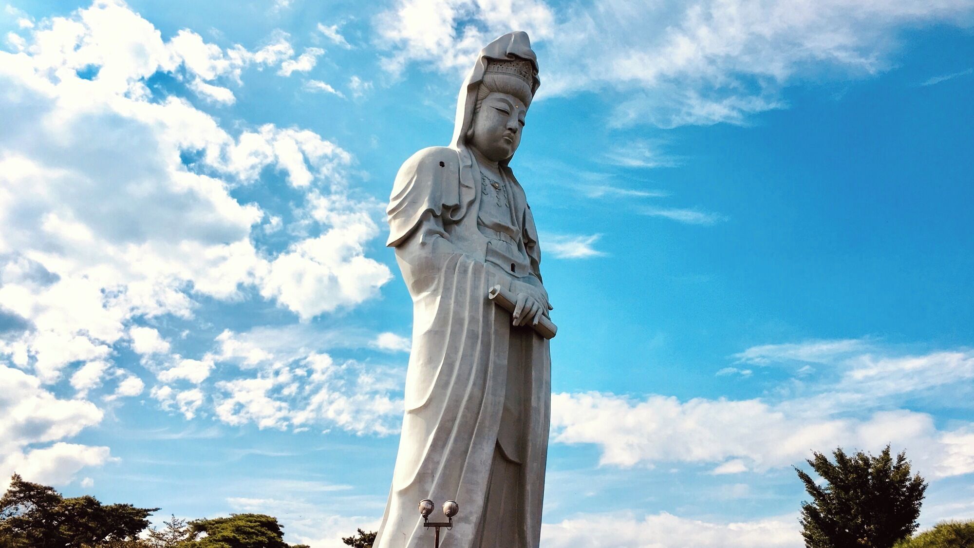A large white statue of a figure with a long robe, standing against a blue sky with clouds.