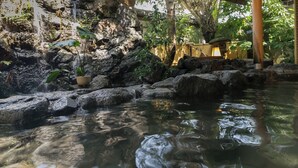 Ein friedliches, überdachtes Wasserelement mit Felsformation und kleinem Wasserfall, umgeben von üppiger Vegetation und einer traditionellen Bambuskonstruktion.