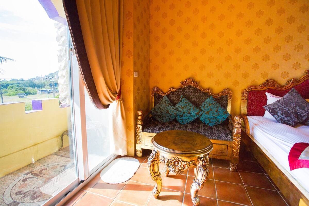 Una habitación con cama, sofá y una pequeña mesa. Hay una ventana con cortinas, y el papel tapiz tiene un diseño estampado.