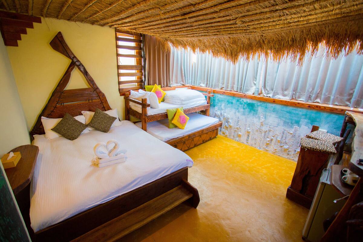 Una habitación con dos camas de madera, una mesita de madera y vistas al mar a través de una ventana con cortinas.