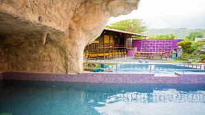 A pool area with a rock formation, a purple building with a red roof, and a view of greenery in the background. - Castillo Kenting (Hengchun)