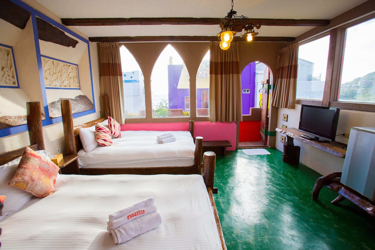 Habitación de hotel con dos camas, una televisión y vistas a edificios a través de ventanales arqueados.