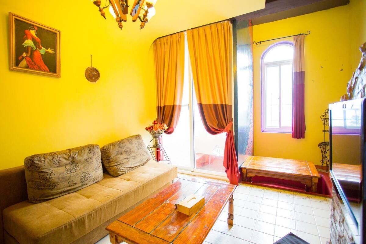Una habitación con sofá, una mesa de centro de madera, un cuadro en la pared y una ventana con cortinas.