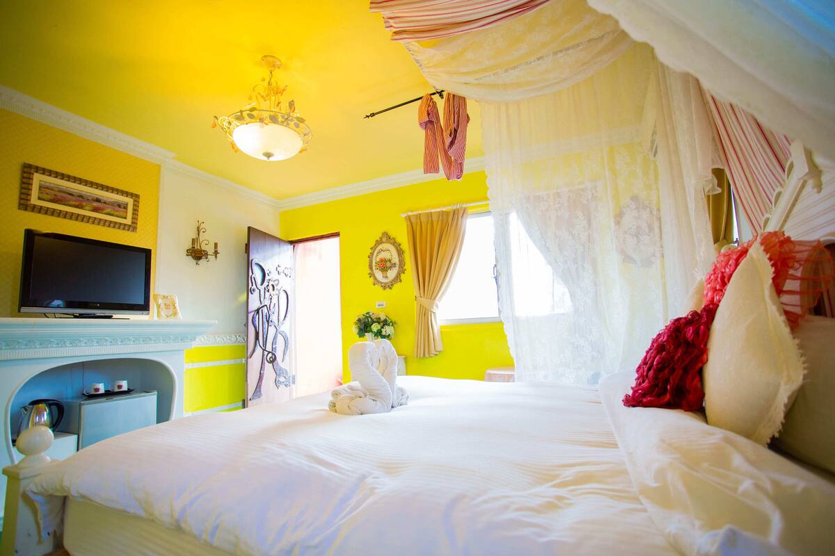 Una habitación luminosa de color amarillo con una cama, un televisor y un escritorio pequeño con una tetera.