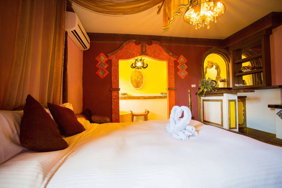 Habitación de hotel con cama, almohadas, una toalla, un candelabro, un equipo de aire acondicionado, un espejo y una pequeña mesa con un florero.