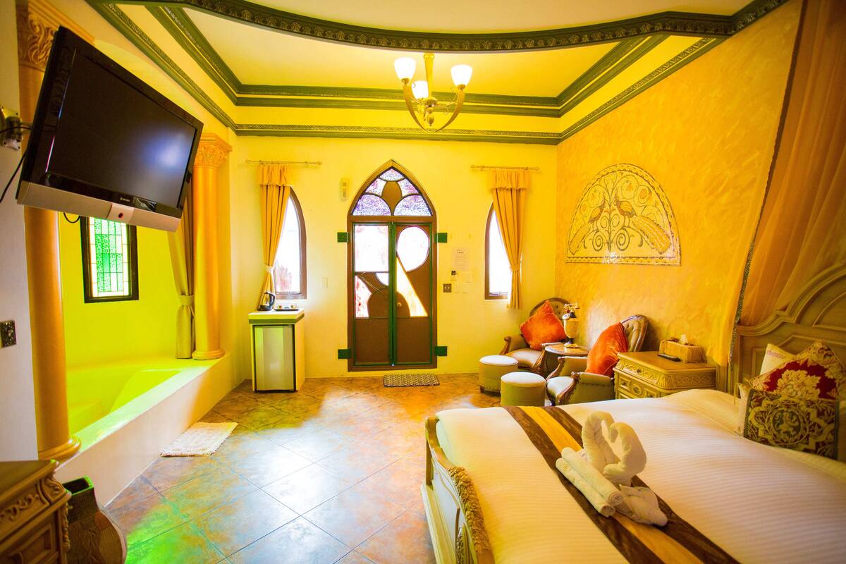 Habitación de hotel con una cama grande, televisor montado en la pared, escritorio pequeño con una silla y ventana con cortinas.