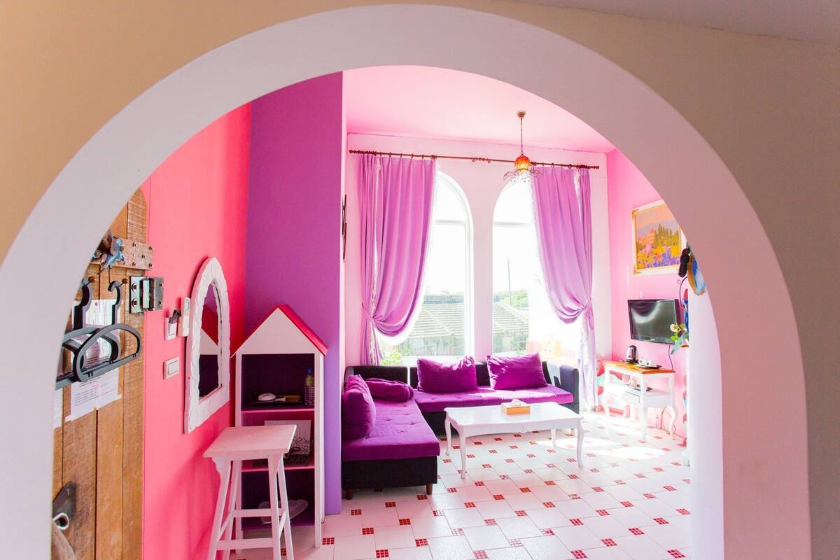 Una habitación de colores vivos con paredes rosas, una mesa de centro blanca, sofás morados y un escritorio pequeño y blanco.