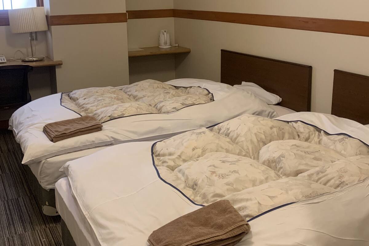 Una habitación de hotel con dos camas, una mesita de noche con lámpara y un pequeño estante con tetera.