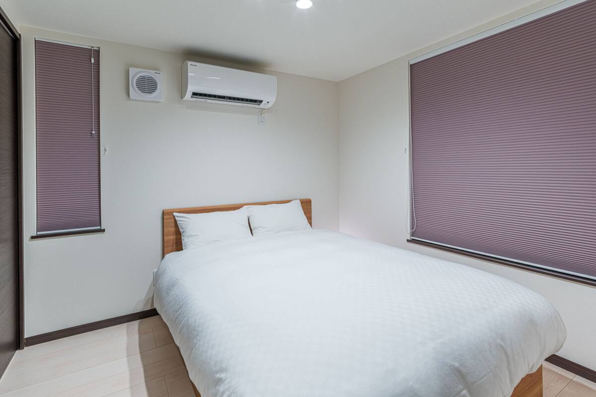 A bedroom with a bed, two air conditioning units, and large windows with blinds.