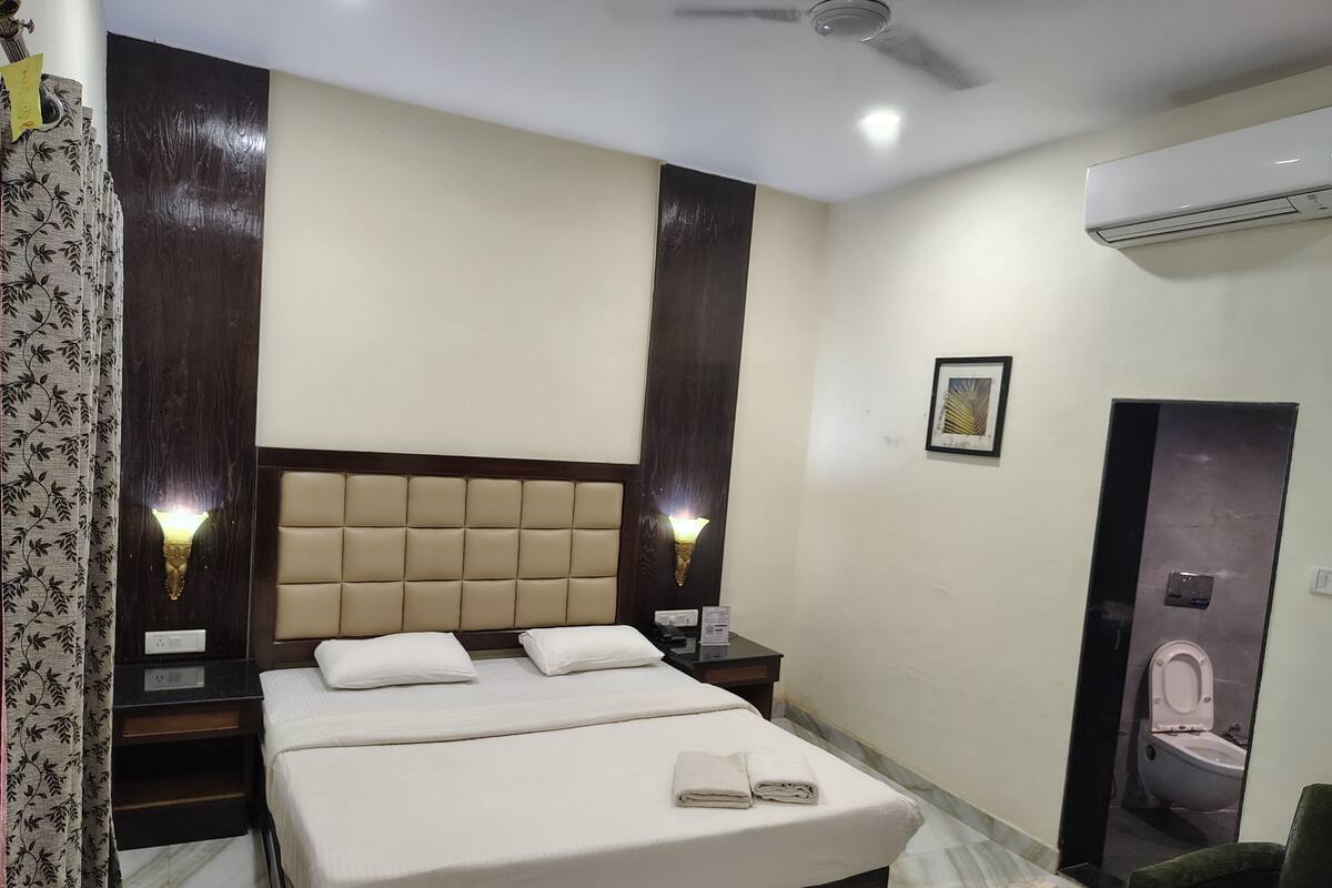 A hotel room with a large bed, two bedside lamps, an air conditioning unit, a framed picture on the wall, and a bathroom door.