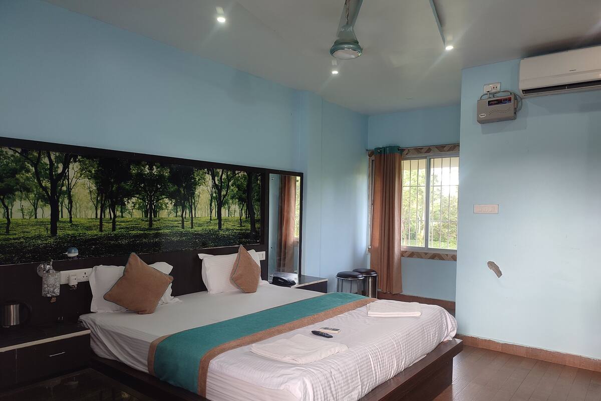 Quarto de hotel com uma cama grande, uma mesa de cabeceira, um espelho, uma janela com cortinas e um aparelho de ar condicionado na parede.
