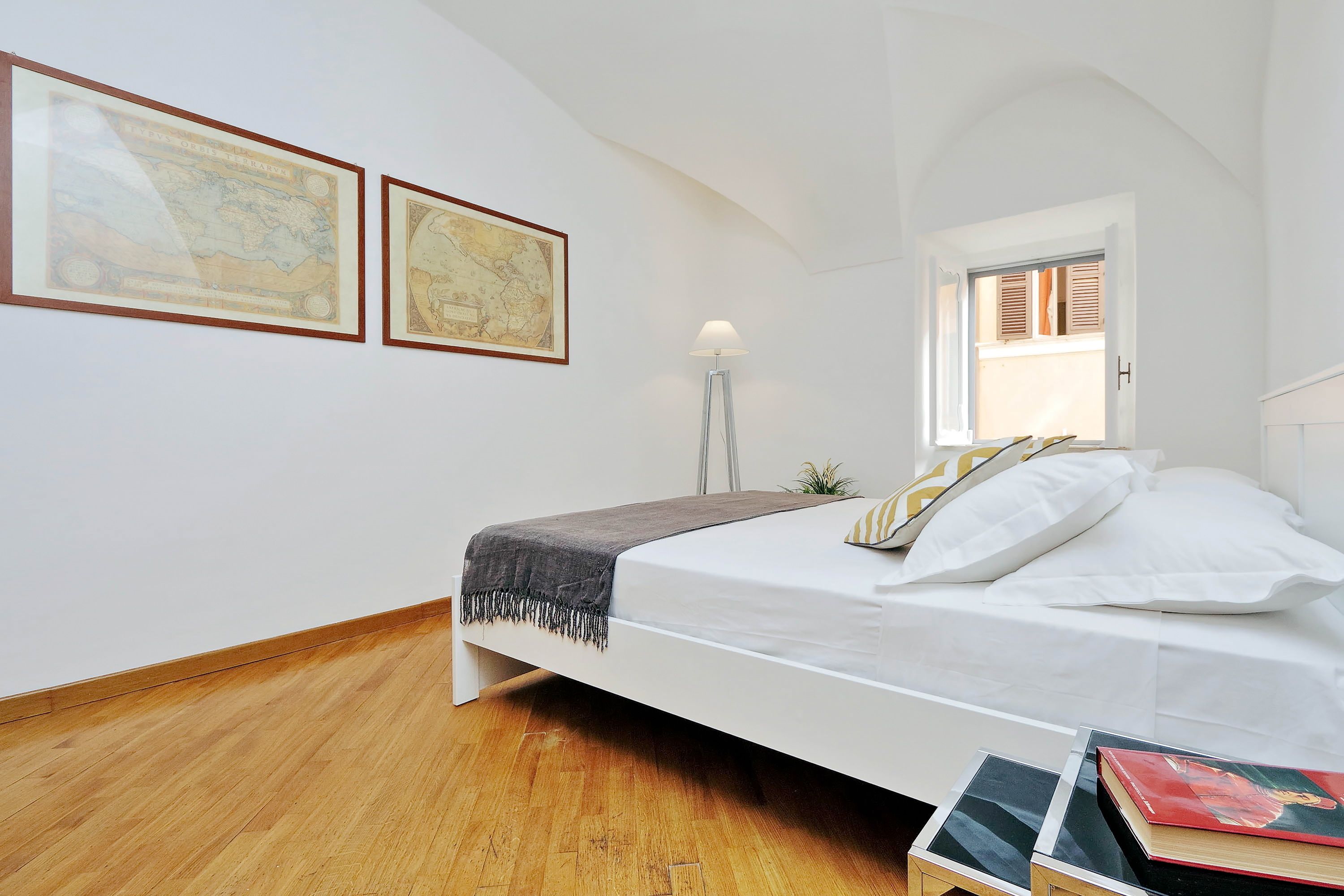 Une chambre à coucher avec un lit, un plancher en bois, deux cartes encadrées accrochées au mur, une lampe sur pied et une fenêtre avec des volets.