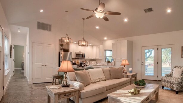 A spacious living room with a beige sofa, two side tables with lamps, a ceiling fan, and a view of the kitchen and patio through glass doors. - Lone Star Cabin No. 6 (Fredericksburg)
