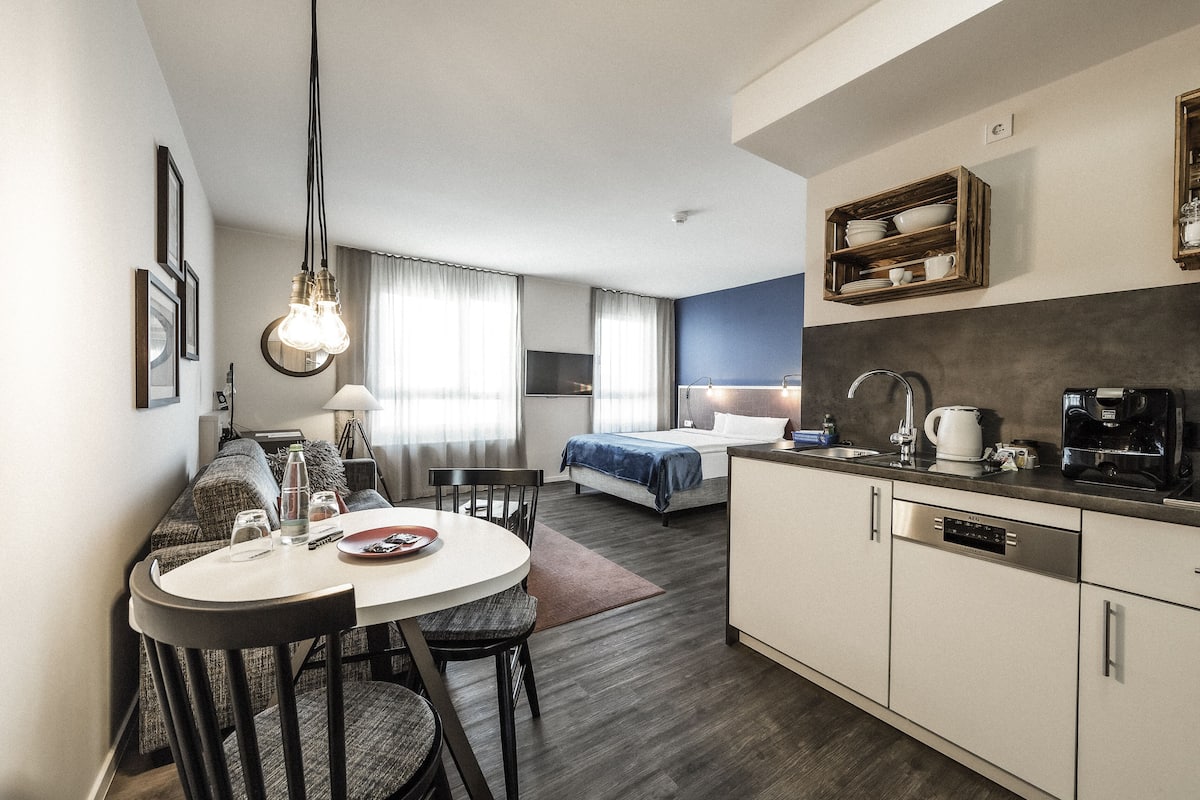 A modern hotel room with a dining area, kitchenette, and a bed with a blue headboard.