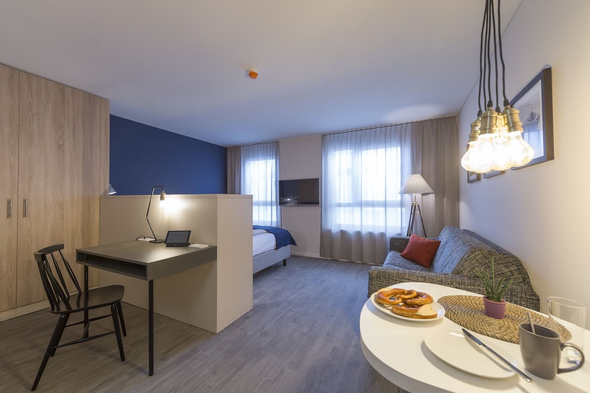 A modern hotel room with a desk, a bed, a sofa, and a dining area.