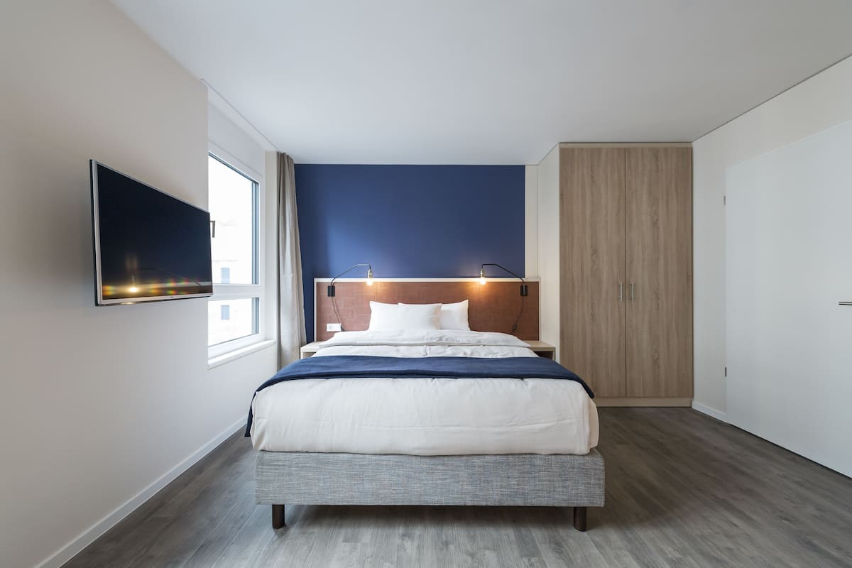 A modern bedroom with a bed, a TV mounted on the wall, and a window.