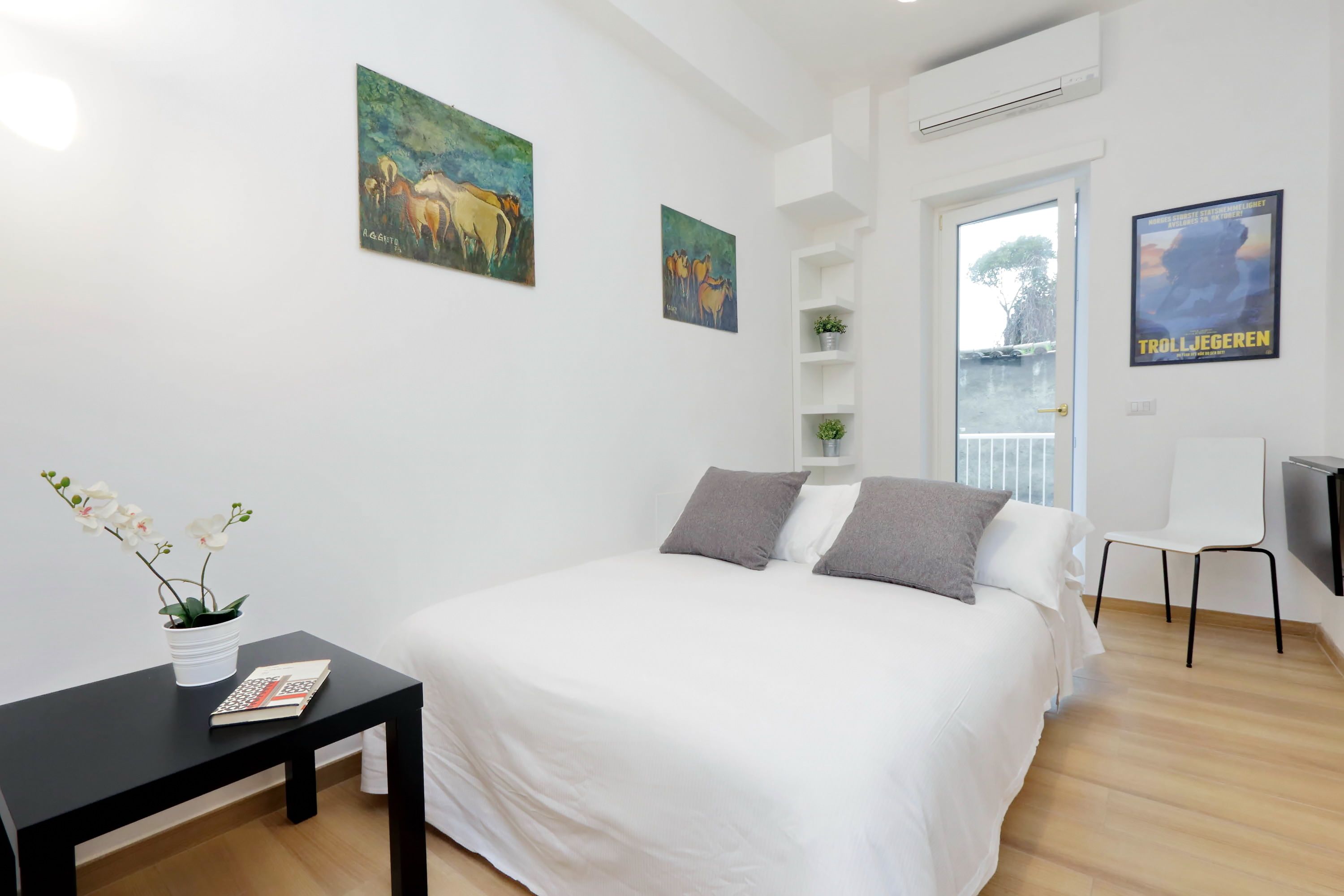 Un dormitorio con cama, mesita de noche, planta en maceta y dos cuadros en la pared.