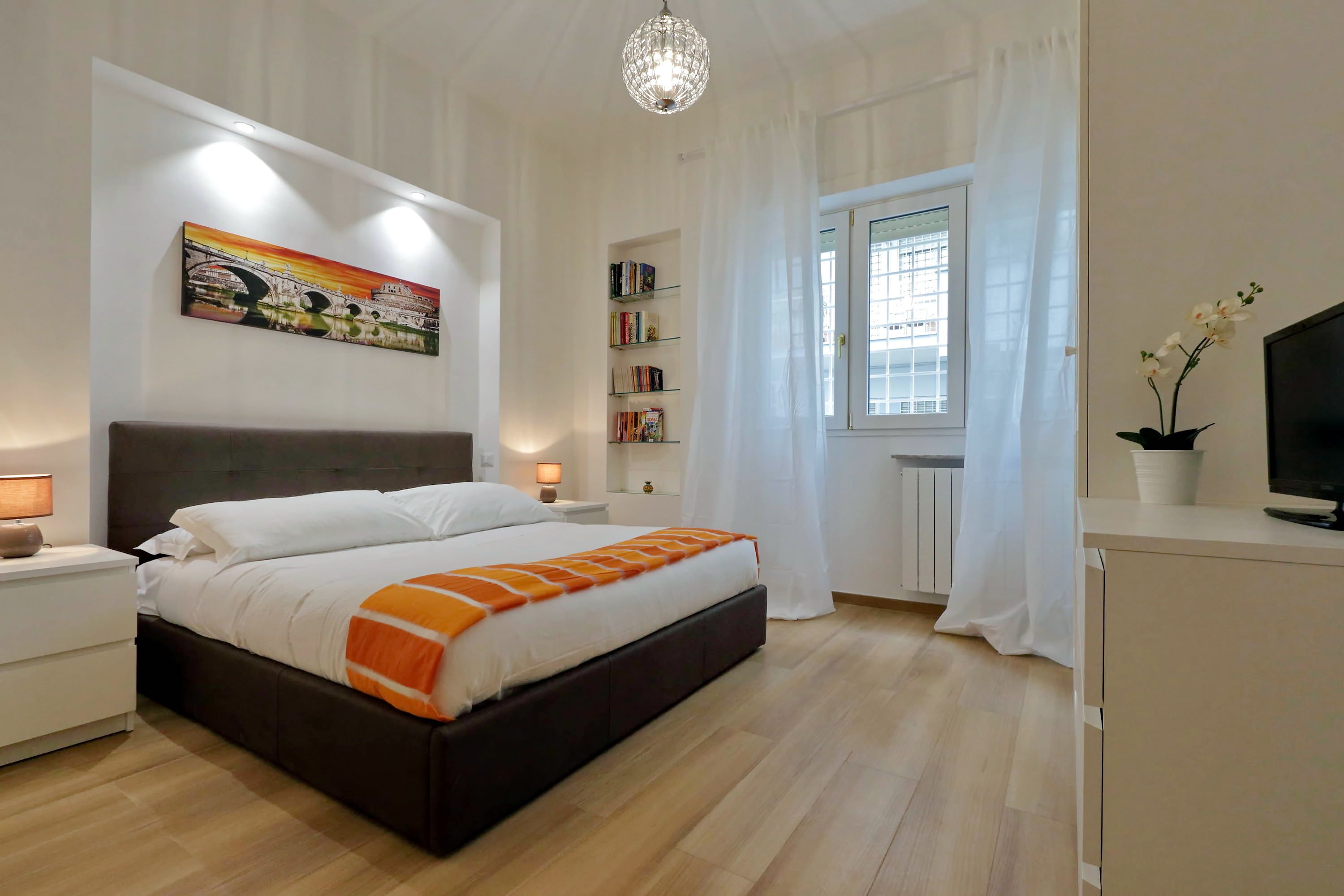 Un dormitorio con cama, mesitas de noche, estantería, ventana con cortinas y televisor.
