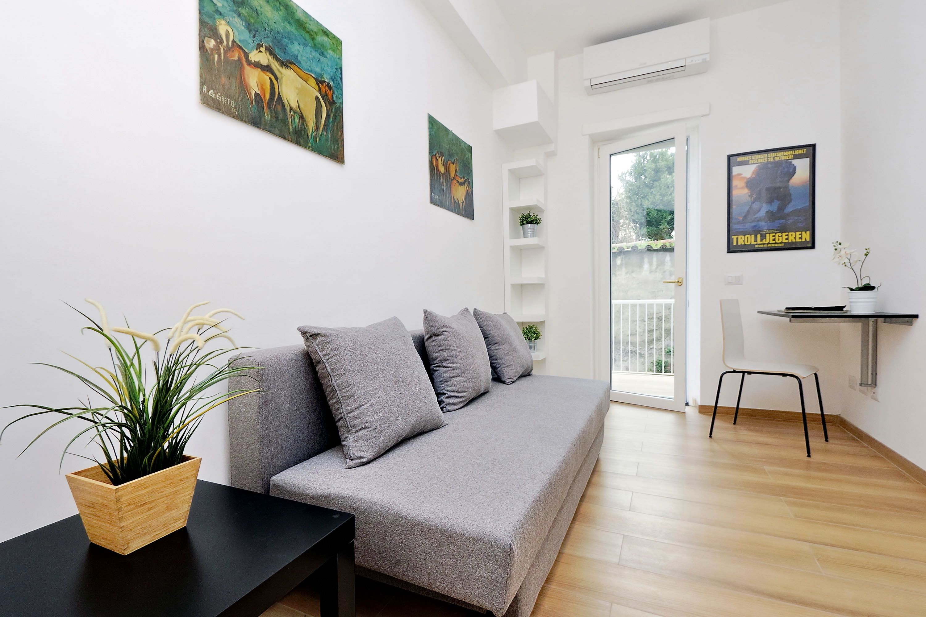 Un salon moderne avec un canapé gris, une table basse noire et une plante en pot. Deux tableaux ornent le mur et une affiche est accrochée à la porte.