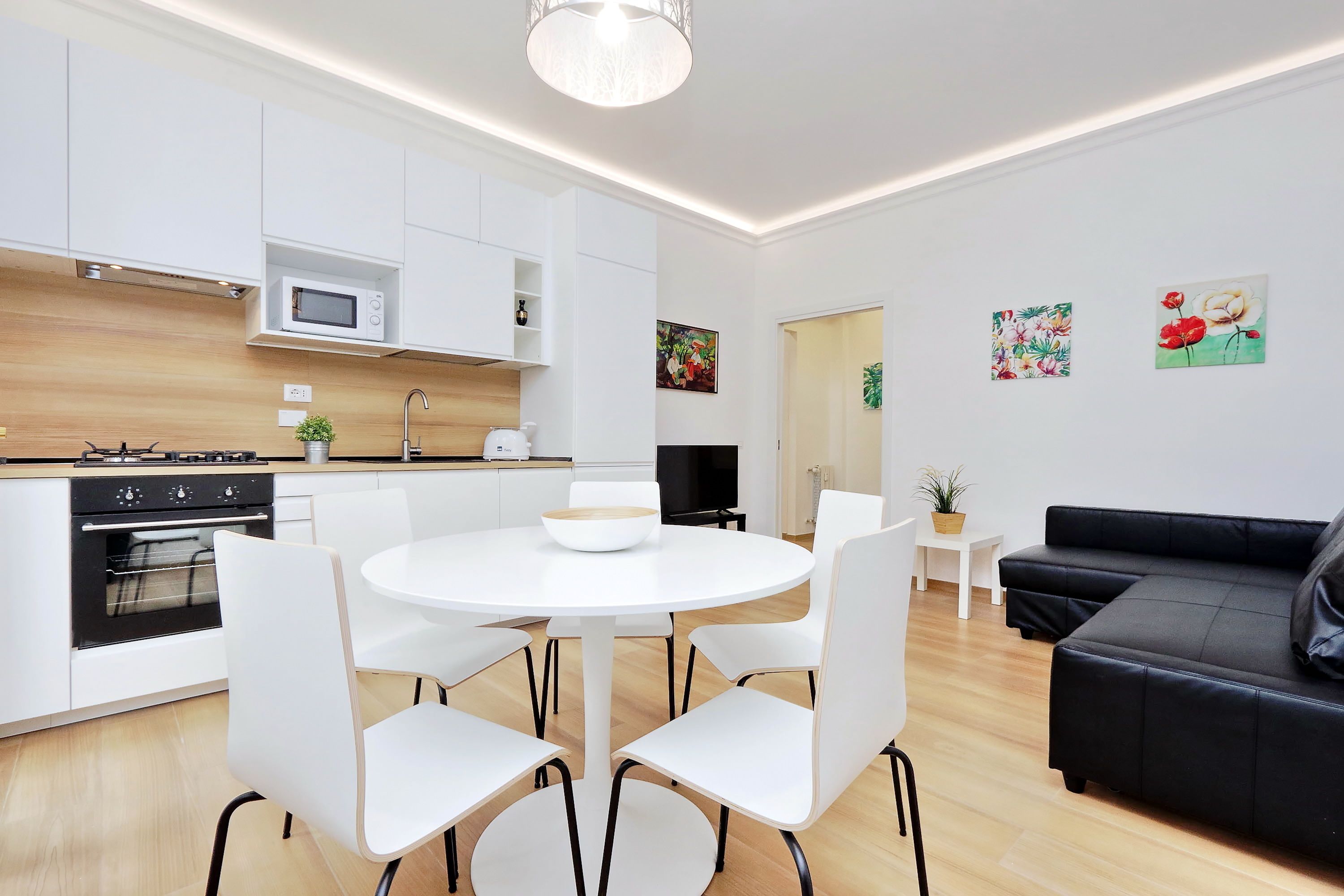 A modern kitchen with white cabinets, a microwave, and a stove. There is a dining area with a white table and chairs. A black sofa is in the living area.