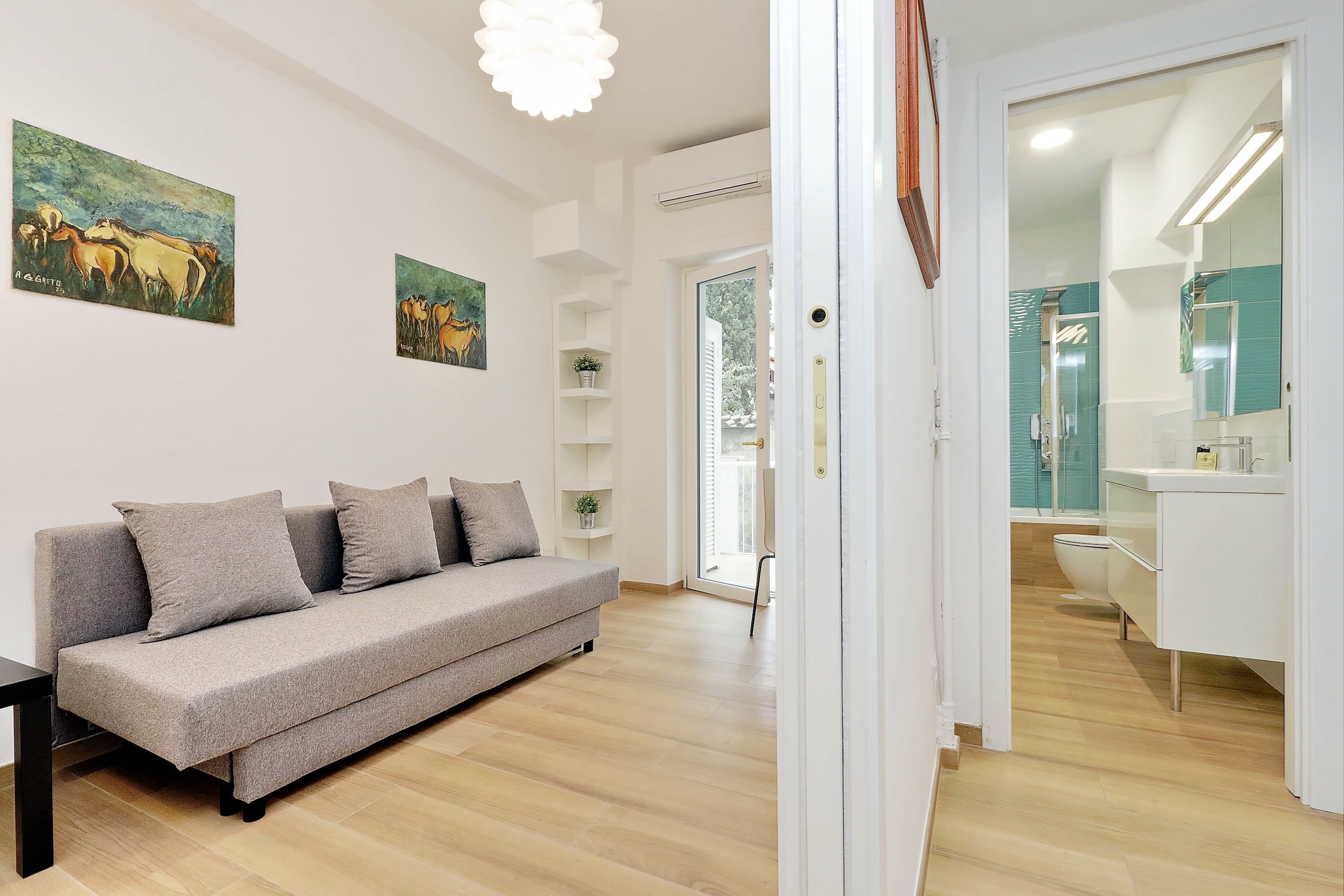 A modern living room with a grey sofa, a coffee table, and two paintings on the wall. There is a door leading to a bathroom with a white sink and mirror.