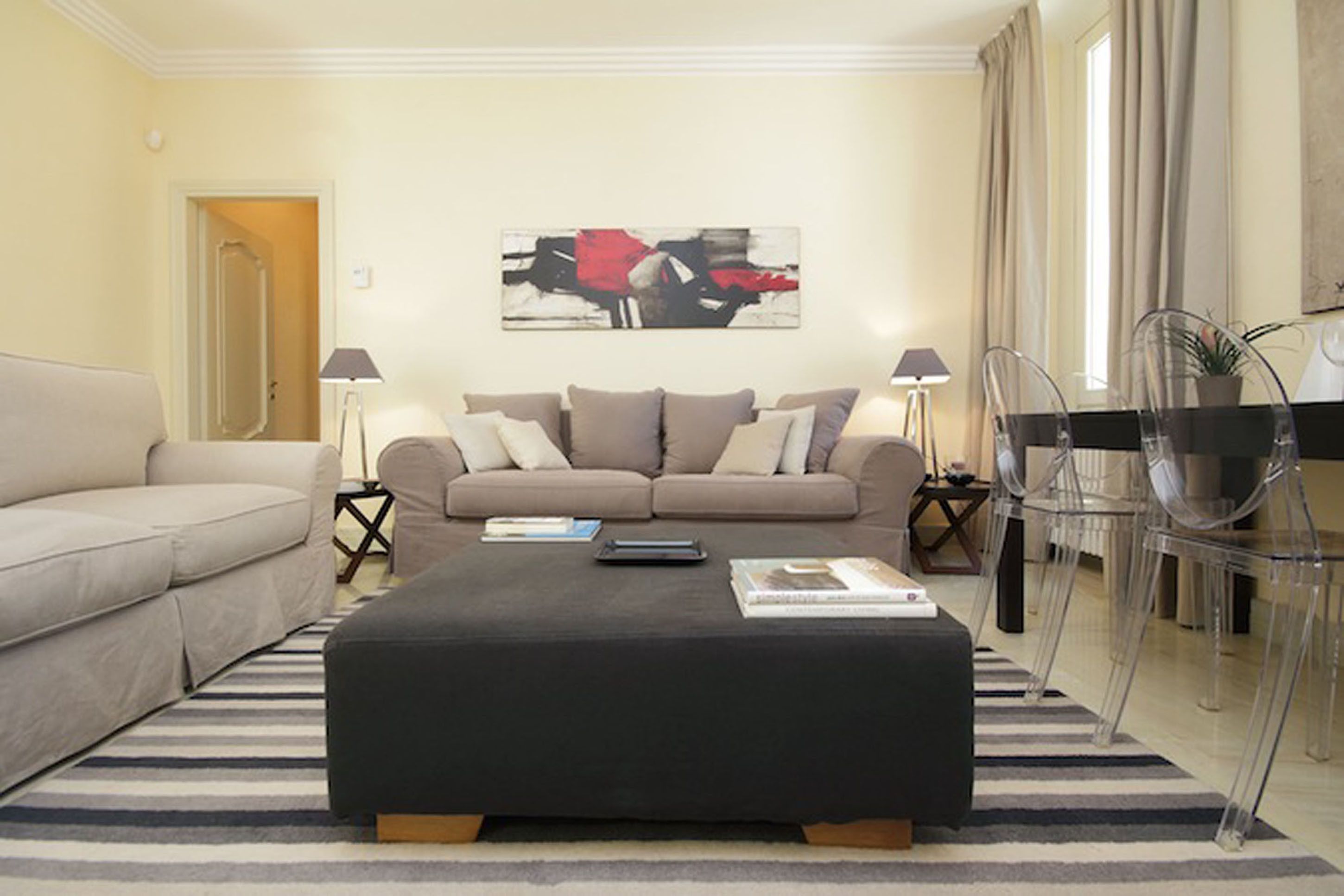A modern living room with a beige sofa, a black ottoman, and a coffee table. There is a painting on the wall, a lamp, and a chair.