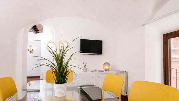 A modern dining area with a glass table, yellow chairs, a white vase with a plant, a flat-screen TV, and a white cabinet. - Fiorentini Charme - My Extra Home (Rome)