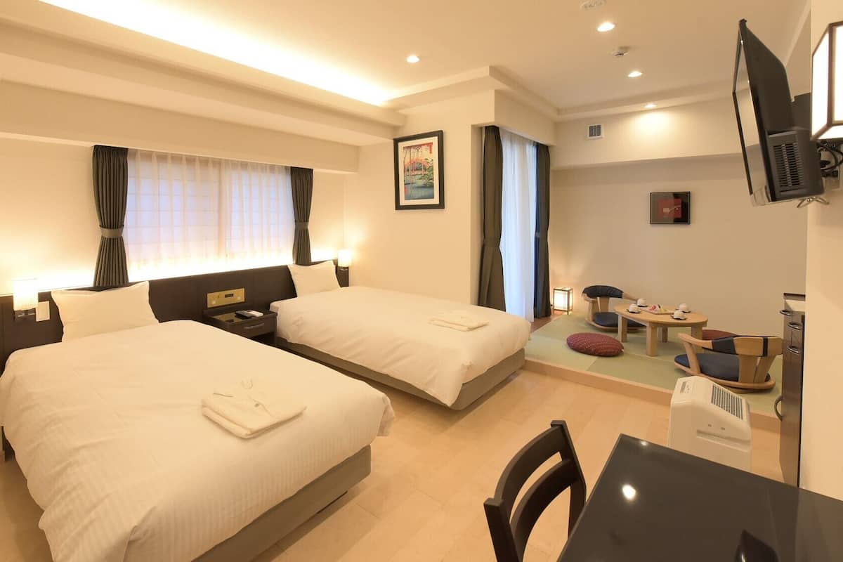 Ein Hotelzimmer mit zwei Betten, einem Schreibtisch, einem Stuhl, einem Fernseher und einer kleinen Essecke.