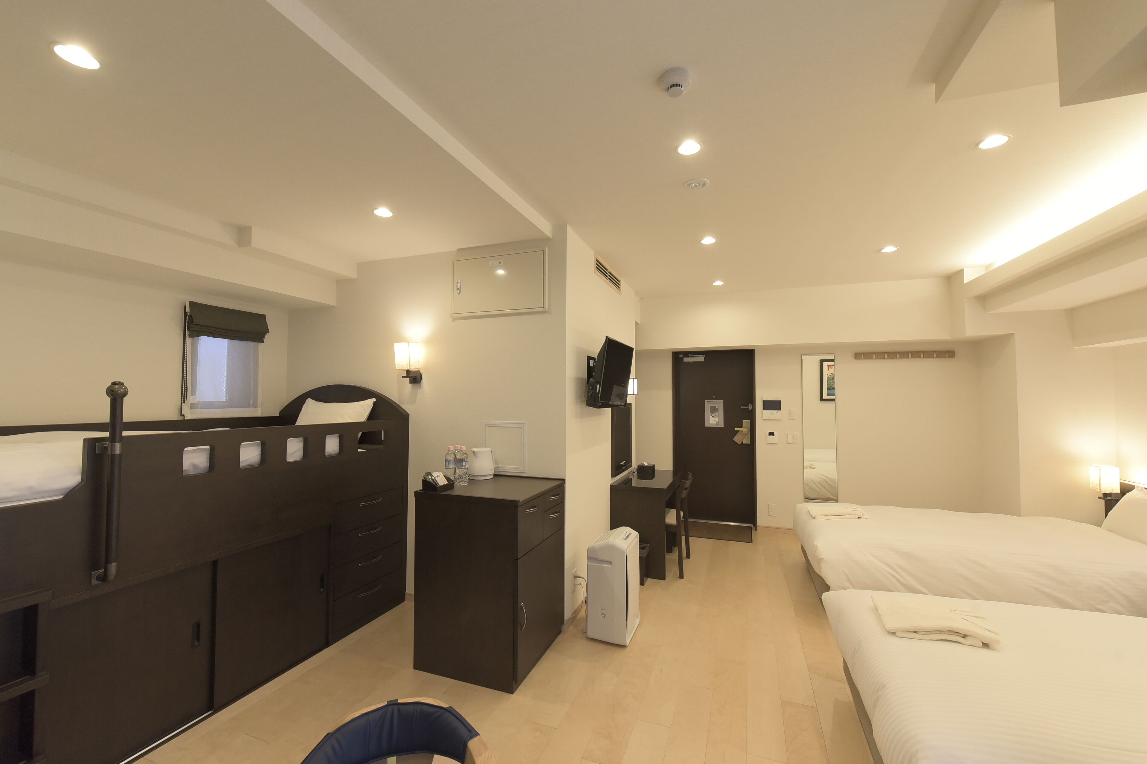A hotel room with a bunk bed, a desk with a chair, a small refrigerator, a bedside table with a lamp, and a flat-screen TV mounted on the wall.