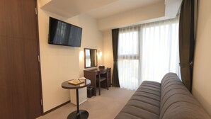 Habitación de hotel con sofá, escritorio, televisión y un amplio ventanal con cortinas.
