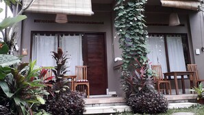 A two-story building with a balcony, a wooden door, and a small garden area with chairs and plants. - Widi House (Ubud)