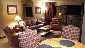 A cozy living room with a sofa, armchairs, a coffee table, and a dining table. There are framed pictures on the wall, a lamp, and a view of the balcony. - Book your Winter get away now (Avon)