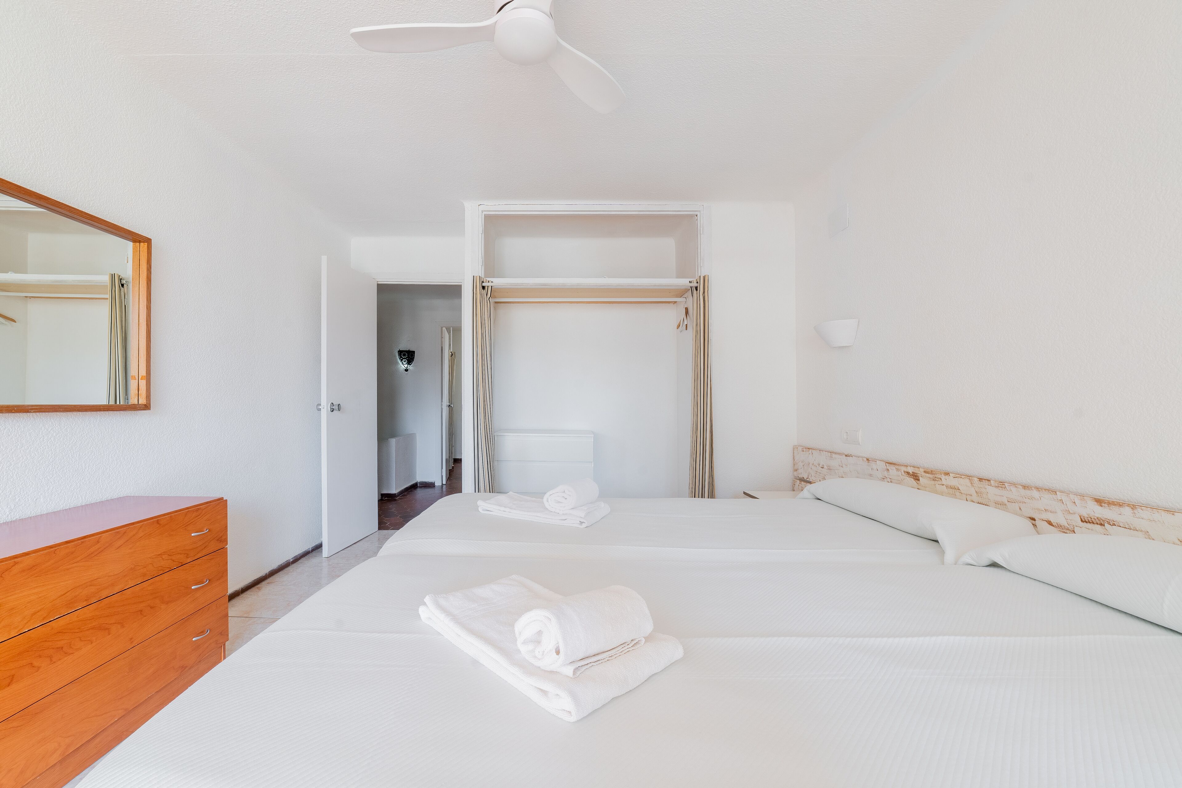 Habitación de hotel con tocador de madera, dos camas con sábanas blancas, ventilador de techo y puerta que da a otra habitación.