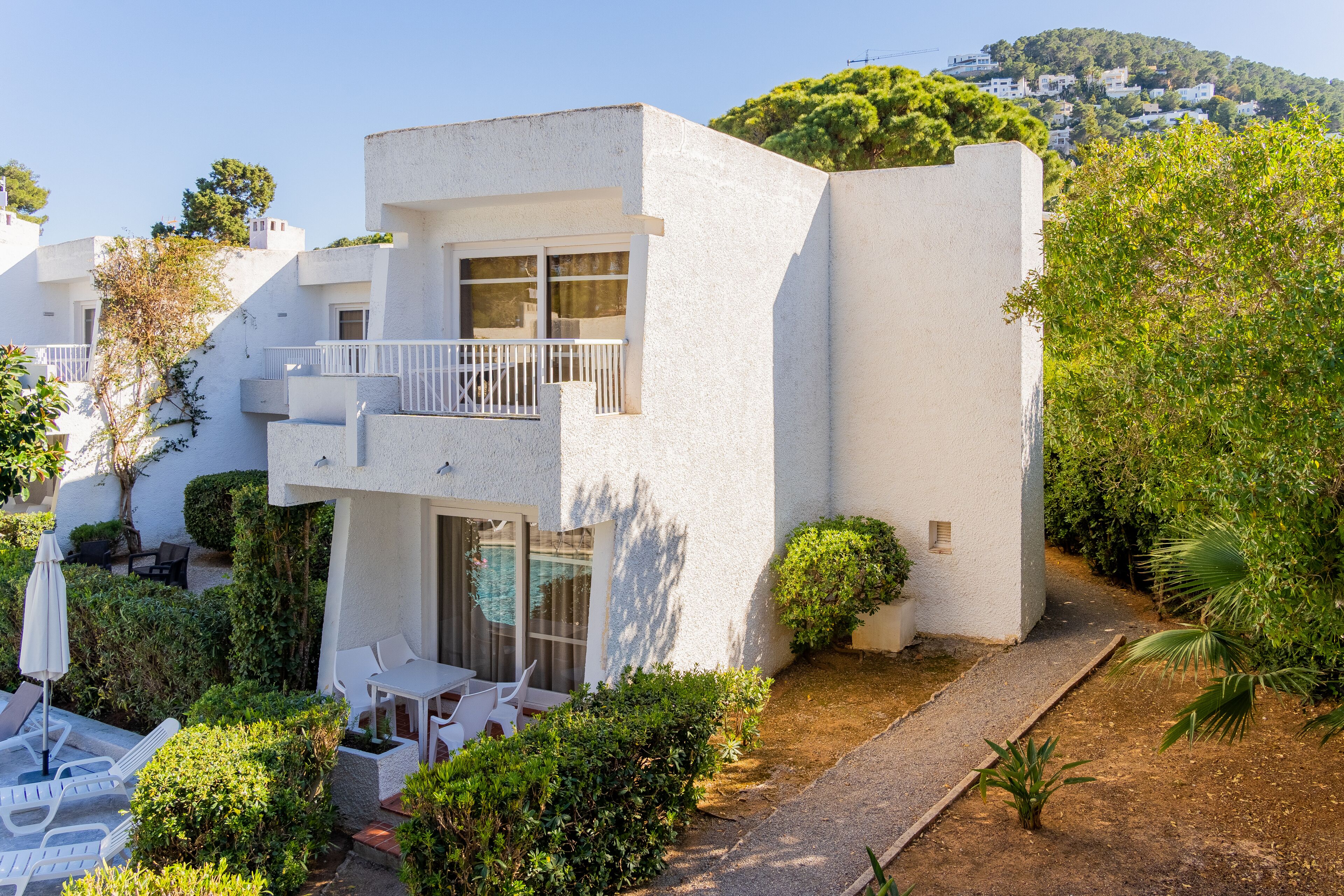 A modern white building with a balcony, surrounded by greenery and a clear sky.