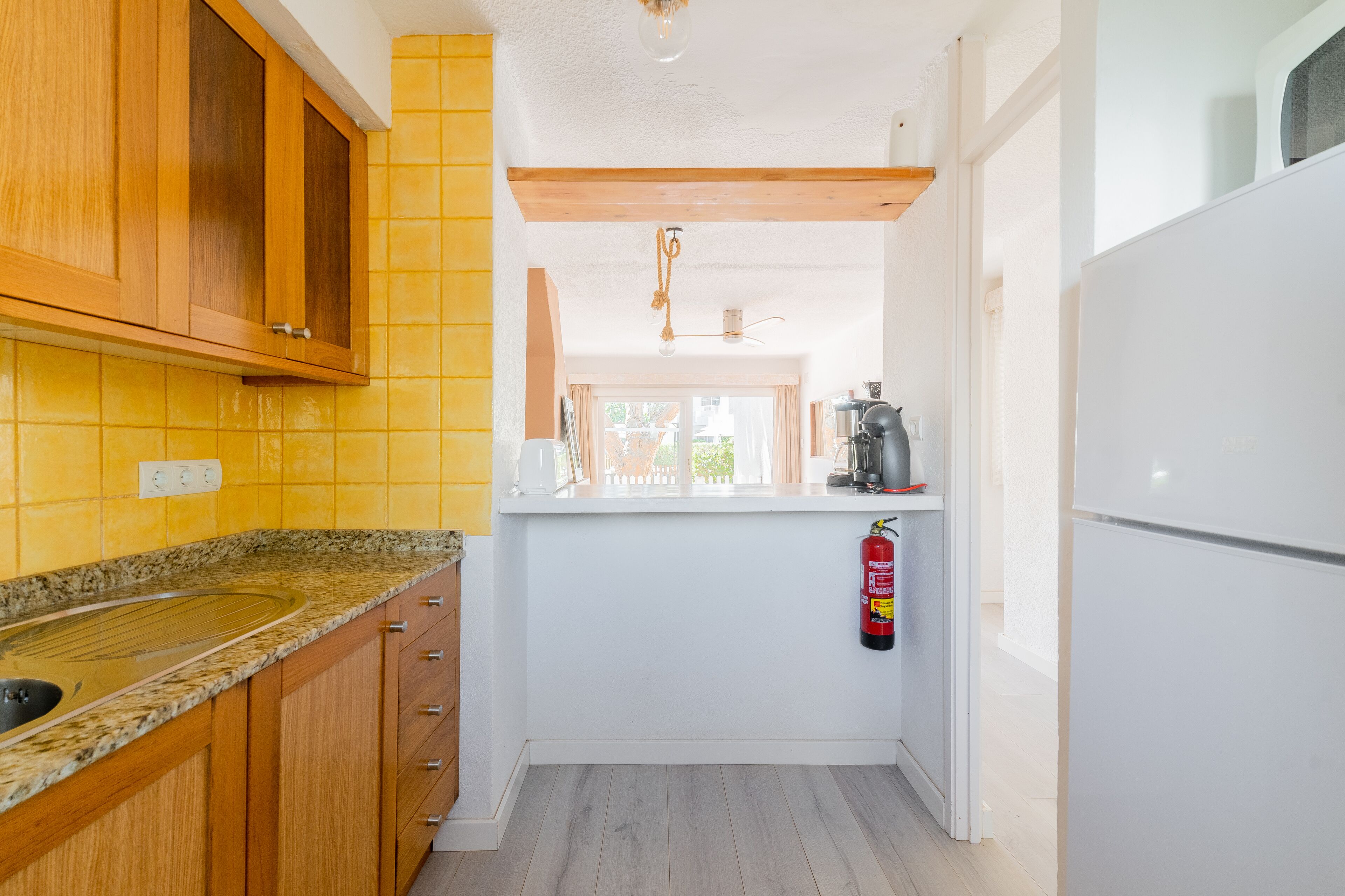 A compact kitchen with wooden cabinets, a granite countertop, a white refrigerator, and a fire extinguisher mounted on the wall.
