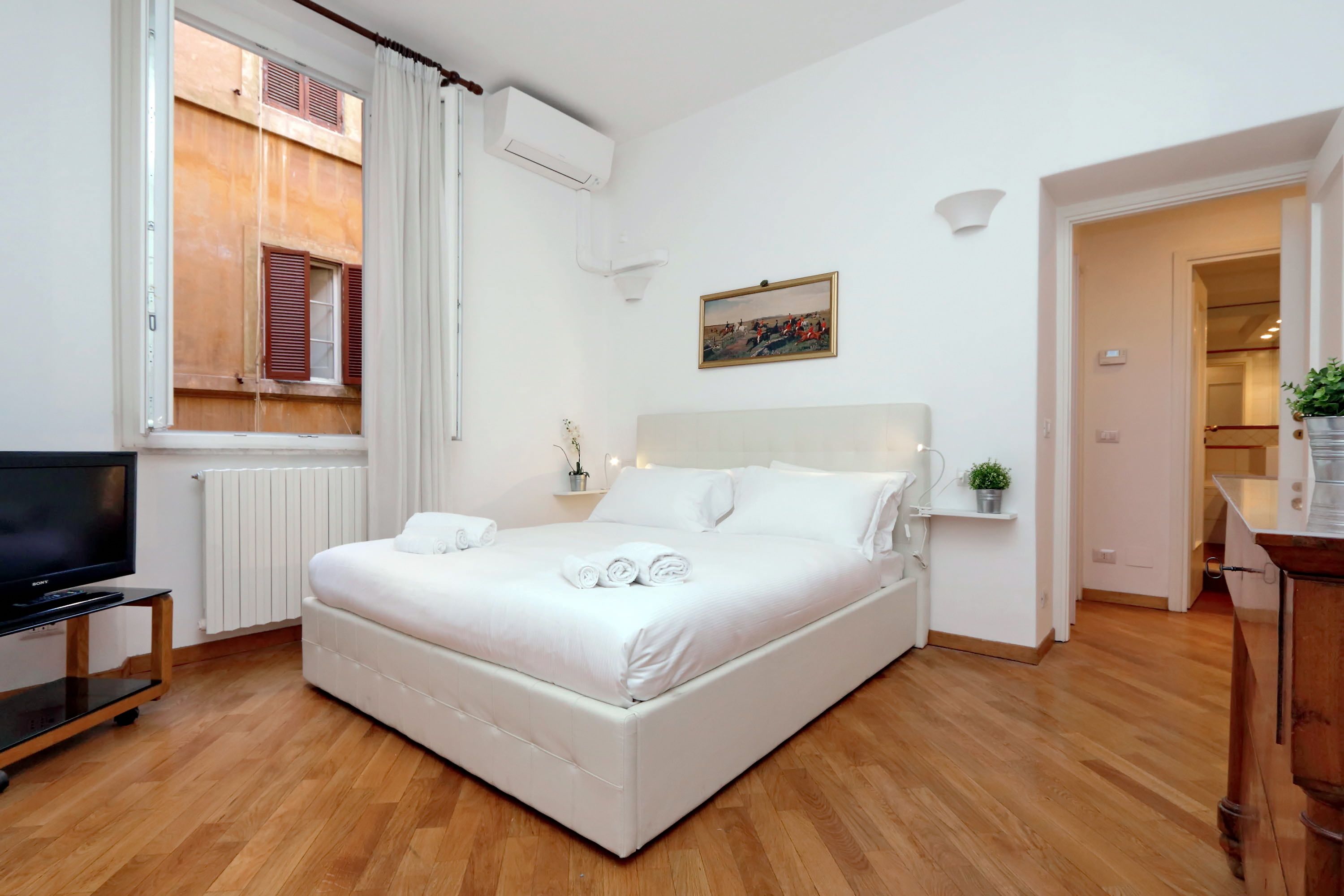 Une chambre bien rangée, avec un lit, un téléviseur posé sur un meuble, une fenêtre avec des volets et un tableau accroché au mur.