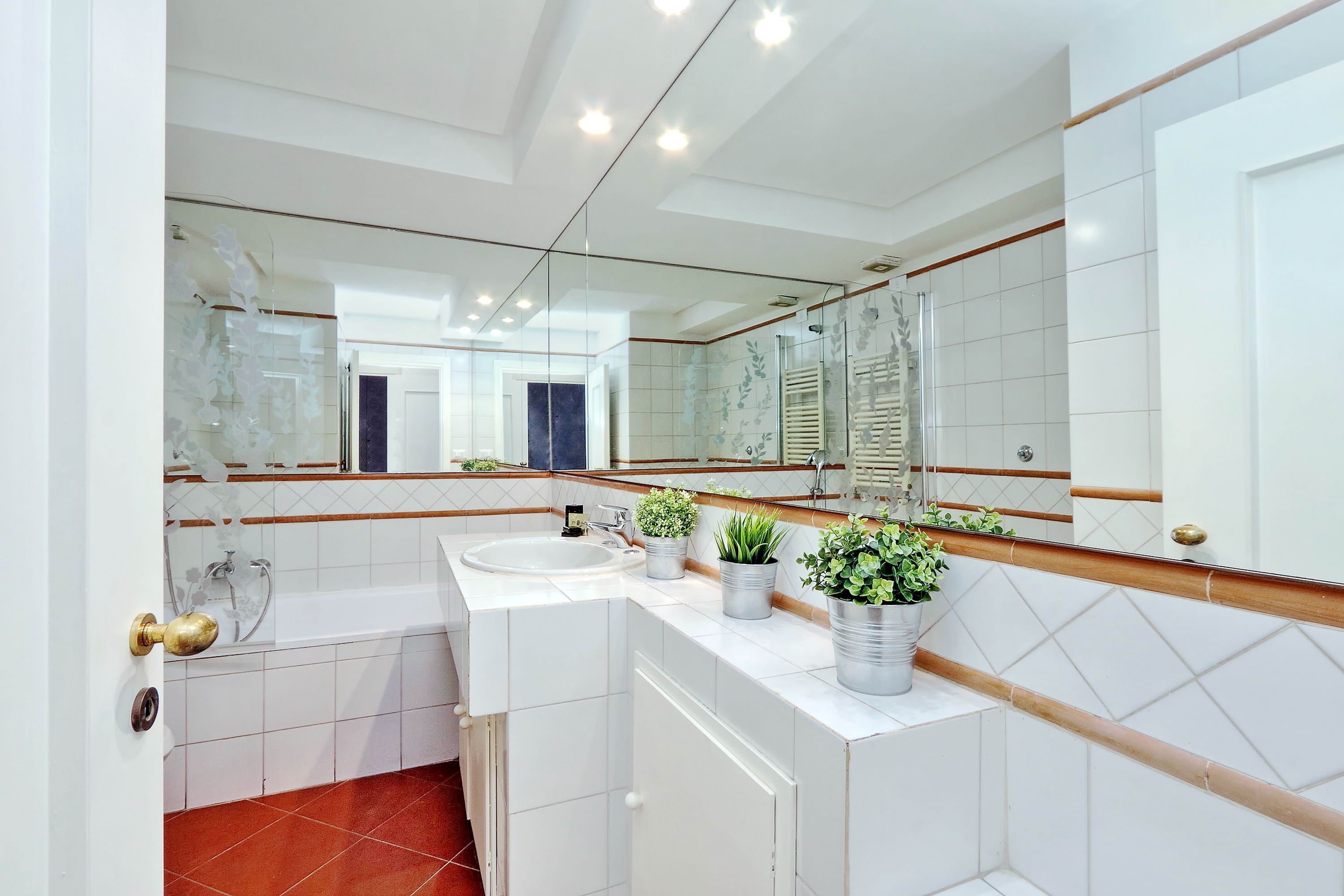 Een moderne badkamer met een grote spiegel, witte tegels, een badkuip en twee potplanten op het aanrecht.