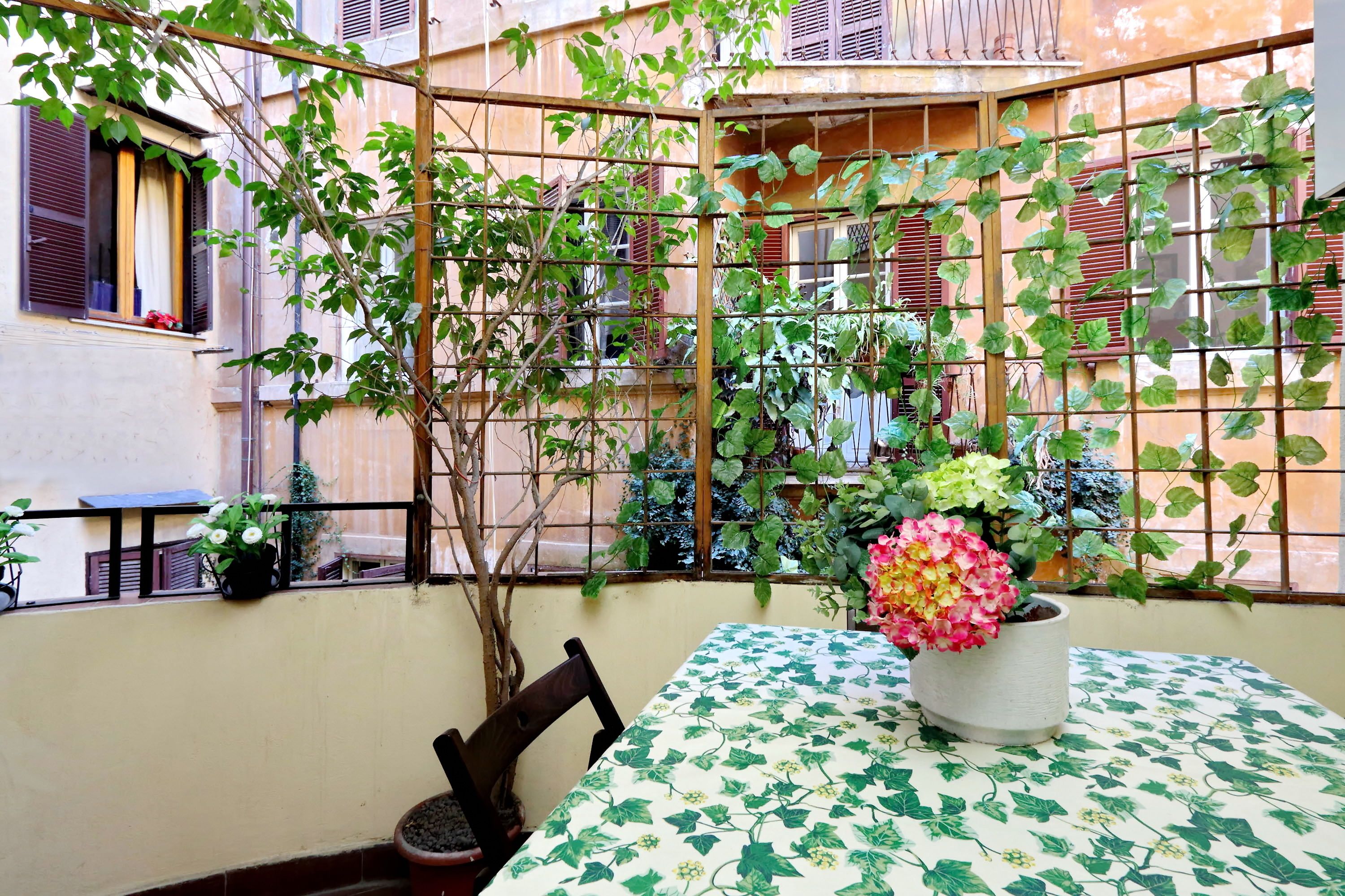 Un balcón con una mesa cubierta por un mantel con estampado de hojas verdes, rodeado de plantas en macetas y una silla de madera.