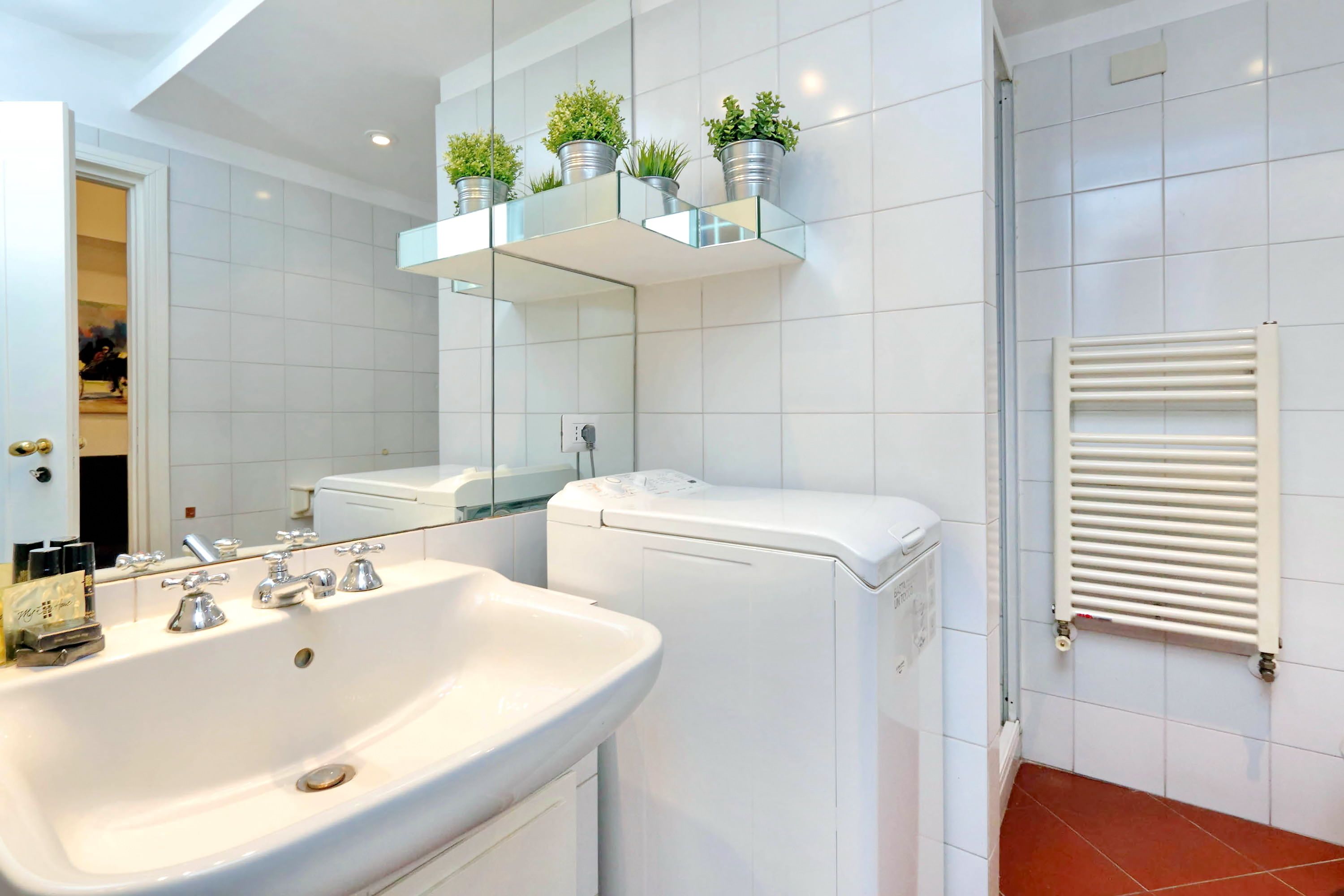 Un baño moderno con un lavabo blanco, un inodoro, un estante de vidrio con plantas y un radiador blanco.