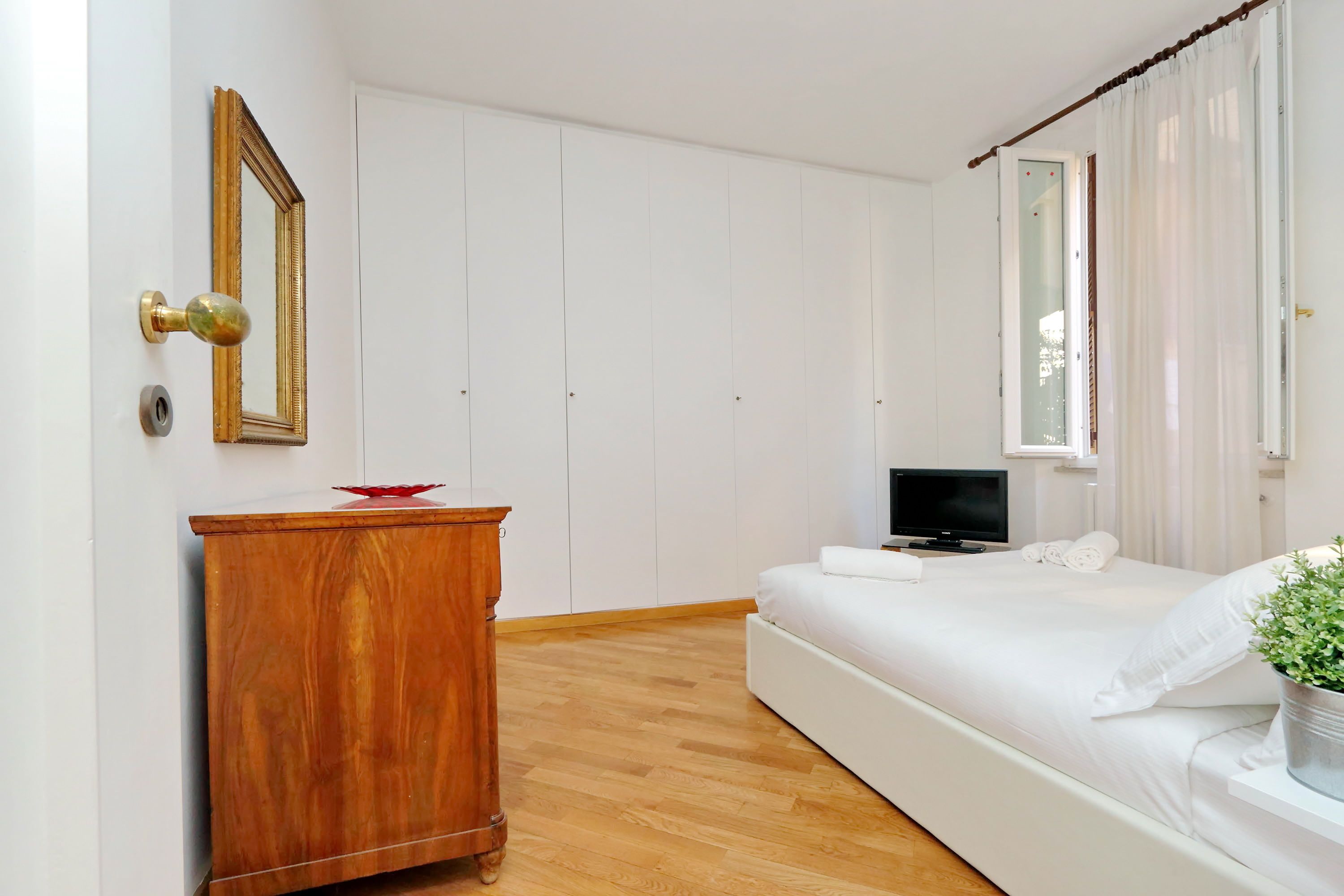 Een slaapkamer met een houten dressoir, een bed met wit beddengoed, een televisie en een raam met gordijnen.